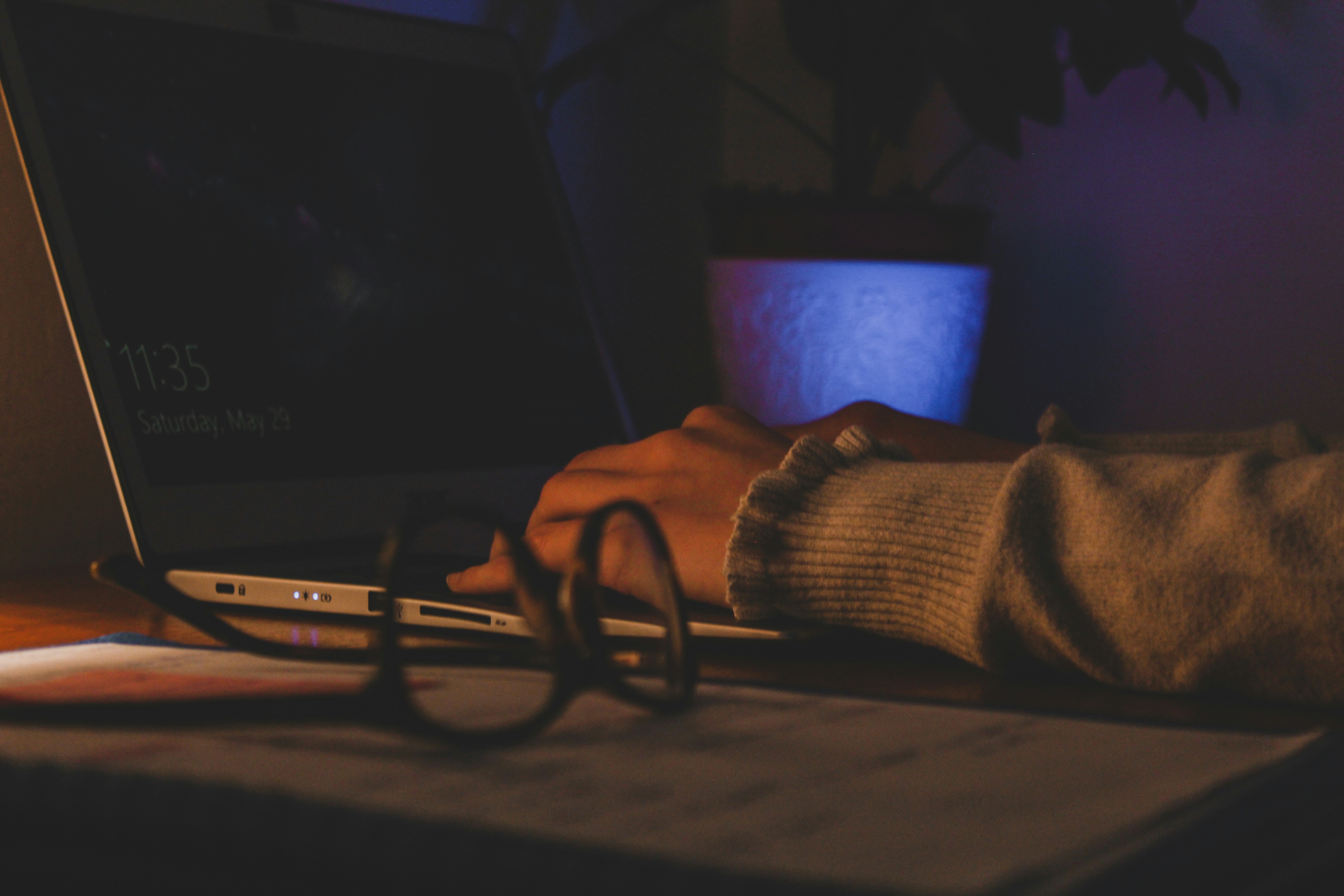 A hand resting on an open laptop in a dark room, the lock screen showing 11:35 on a Saturday night, glasses set down beside it